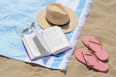 Photo of Beach towel with book, straw hat, sunglasses and flip flops on sand