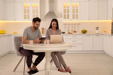 Photo of Happy couple in pajamas using gadgets at kitchen table