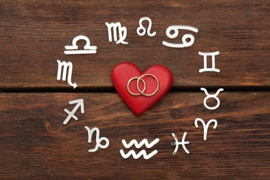 Photo of Zodiac signs, red heart and wedding rings on wooden background, flat lay
