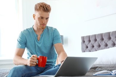 Photo of Young man using laptop while sitting on bed at home