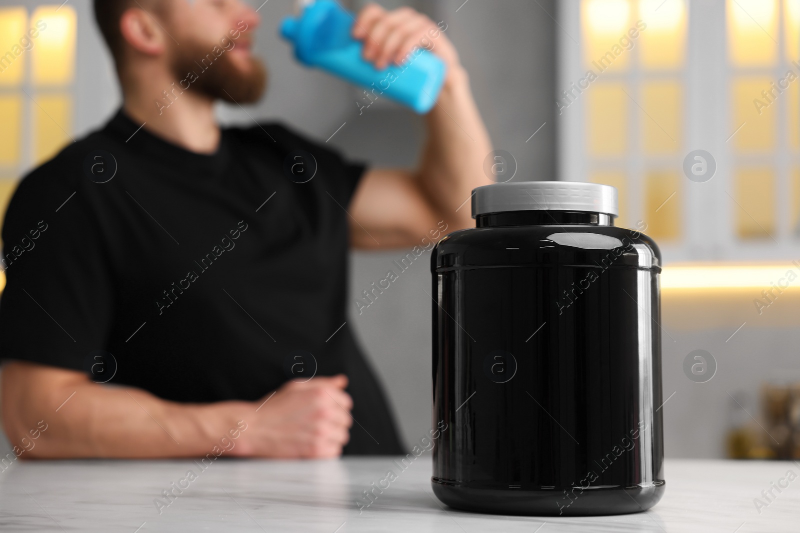 Photo of Young man with shaker of protein at white marble table in kitchen, focus on jar of powder