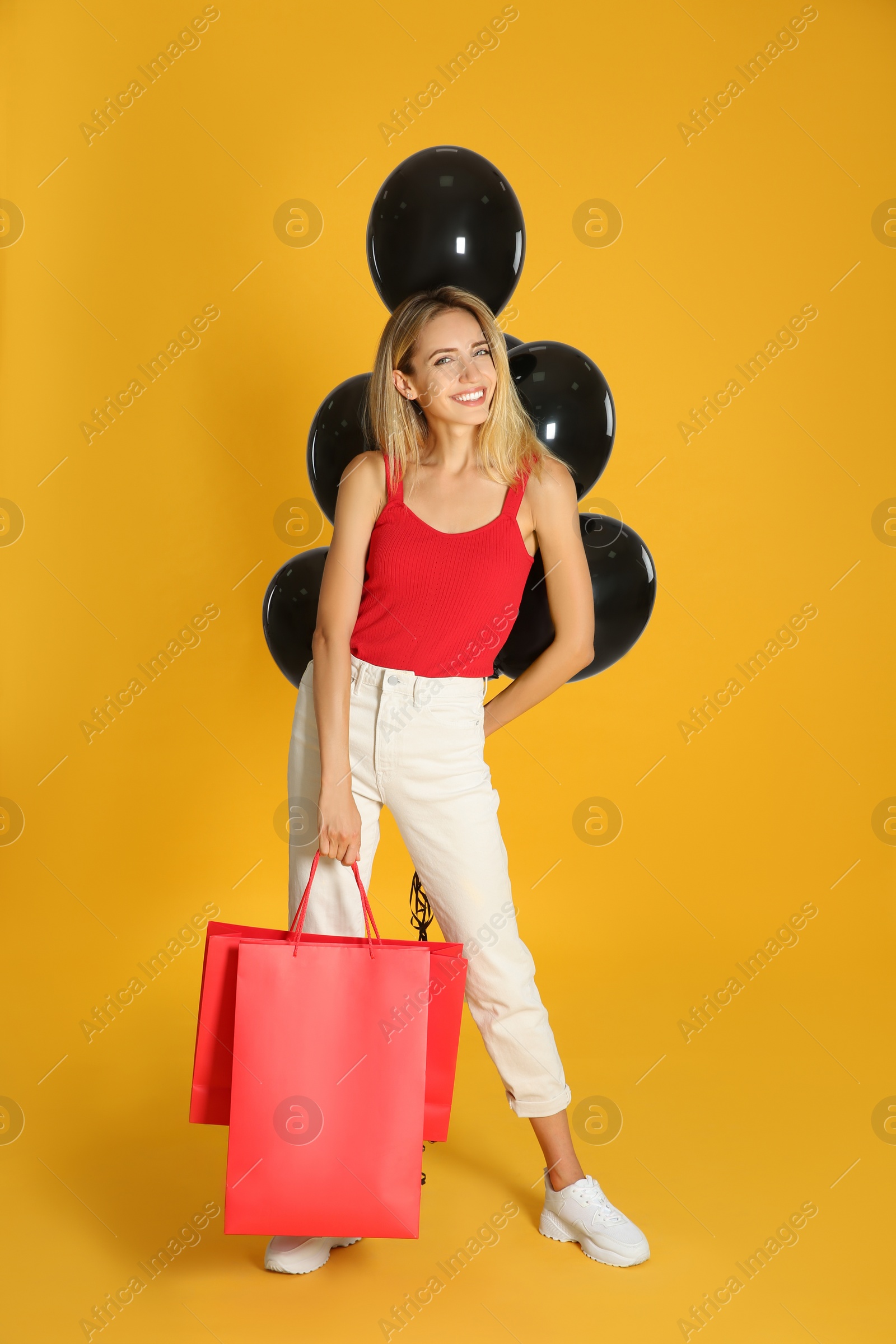 Photo of Happy young woman with balloons and shopping bags on yellow background. Black Friday Sale