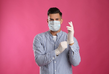 Man in protective face mask putting on medical gloves against pink background