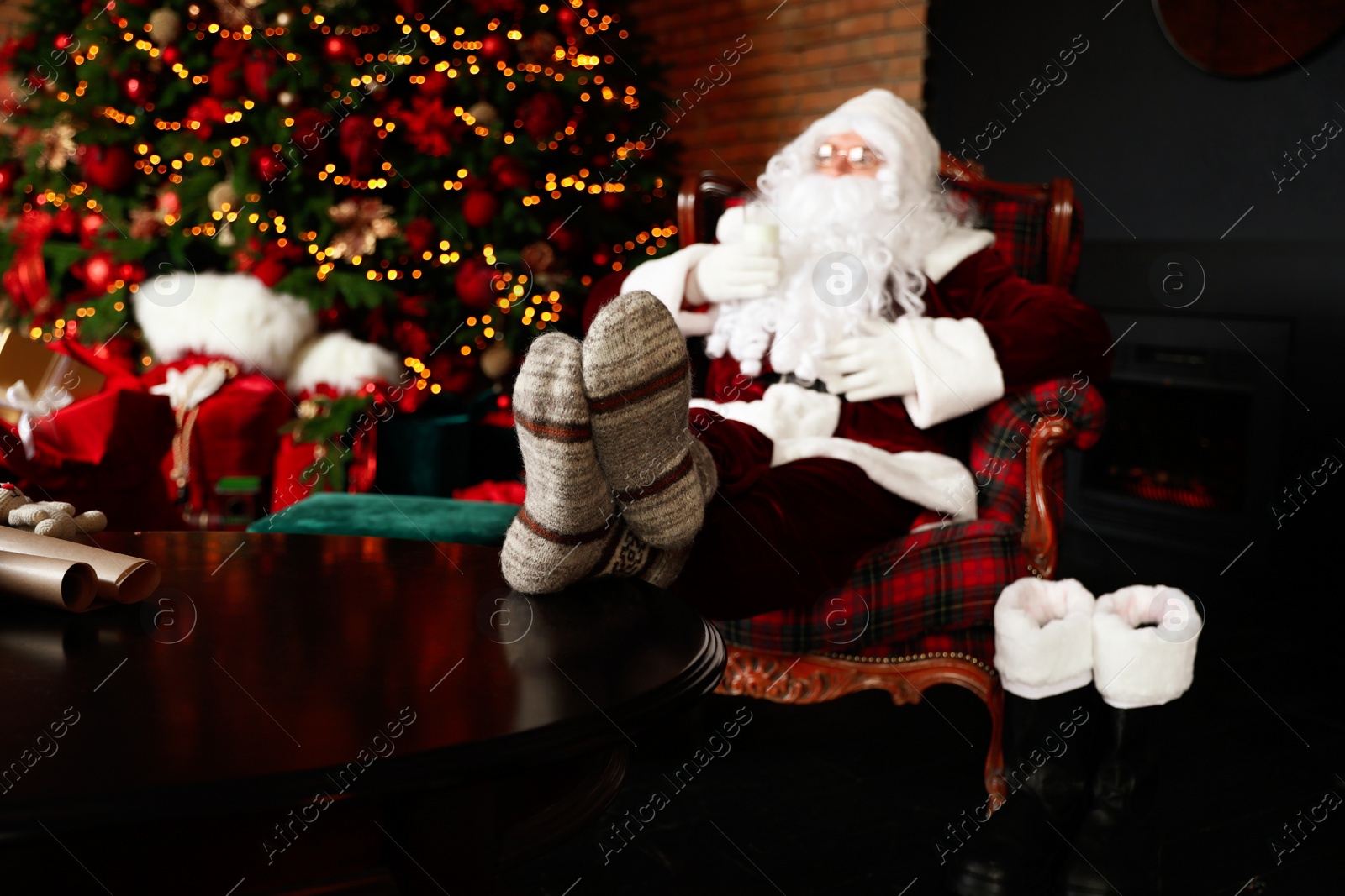 Photo of Santa Claus with glass of milk resting in armchair near Christmas tree, focus on legs