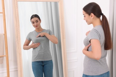 Photo of Beautiful young woman doing breast self-examination near mirror indoors