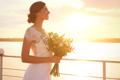 Gorgeous bride in beautiful wedding dress with bouquet near river on sunset