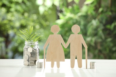 Photo of Pension savings. Figure of senior couple, jar with coins and twig on white table against blurred green background