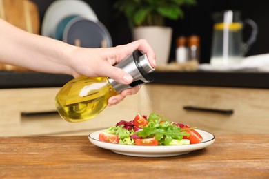 Photo of Woman spraying cooking oil onto salad on wooden table in kitchen, closeup