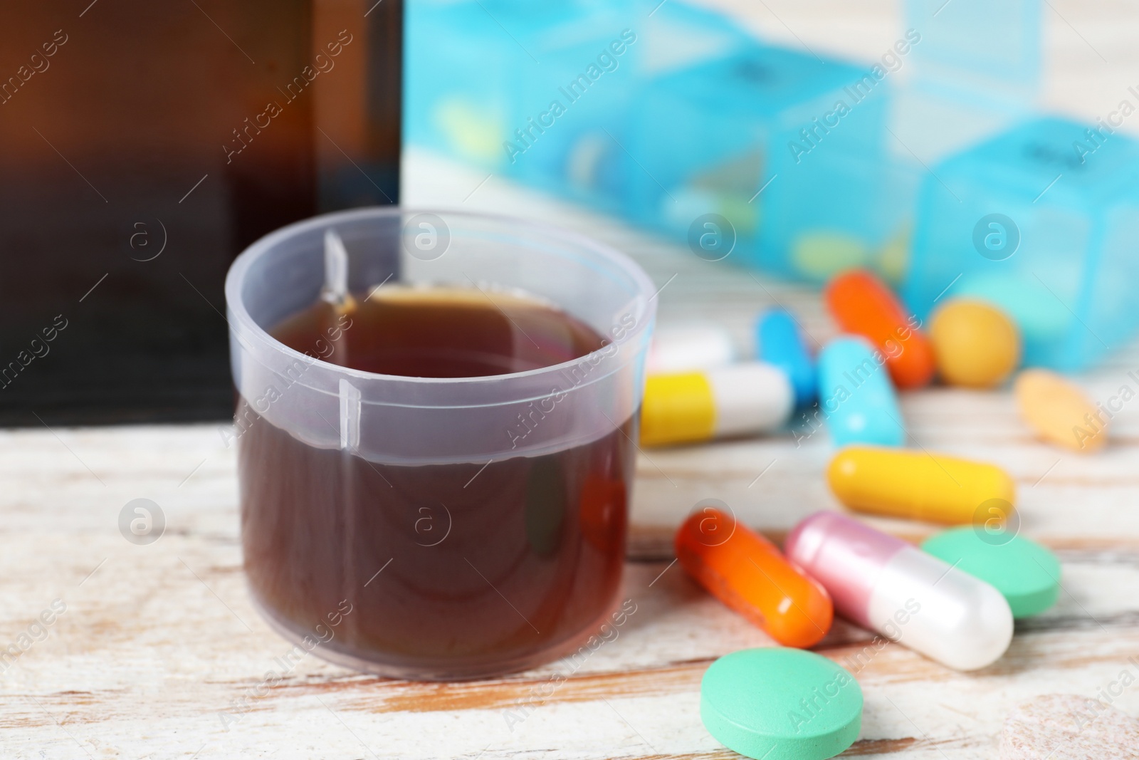 Photo of Measuring cup with syrup and pills on white wooden table, closeup. Cold medicine