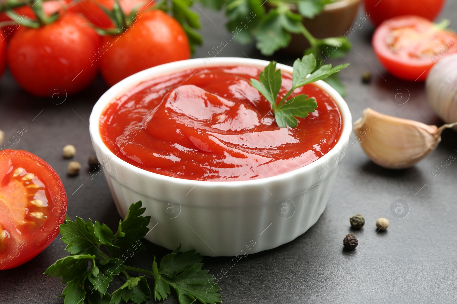 Photo of Delicious ketchup in bowl, tomatoes, parsley and garlic on grey table, closeup