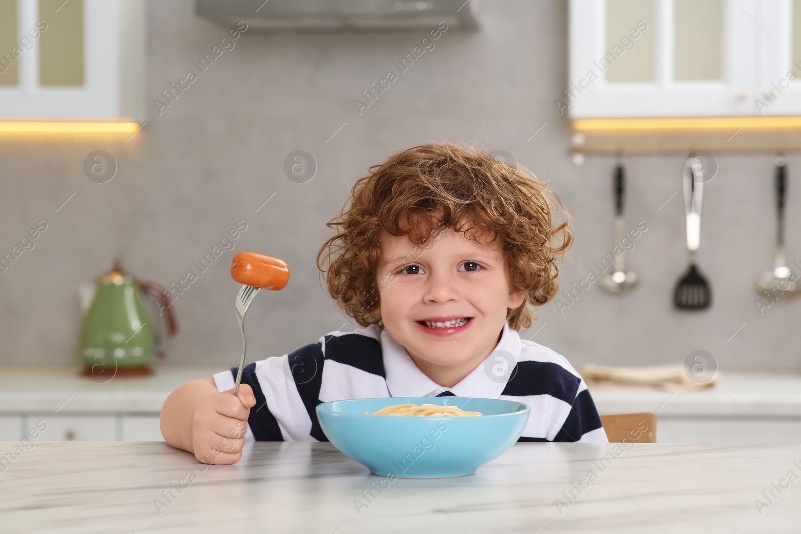 Photo of Cute little boy holding fork with sausage and bowl of pasta at table in kitchen