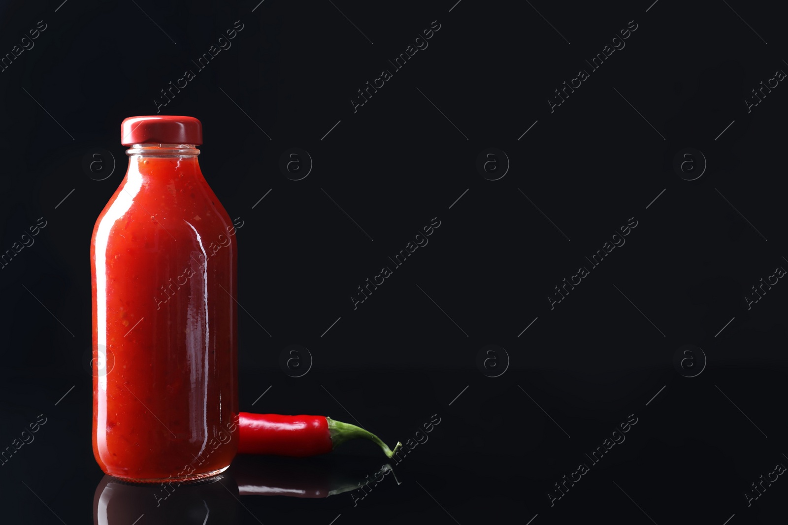 Photo of Spicy chili sauce in bottle and pepper against dark background, space for text