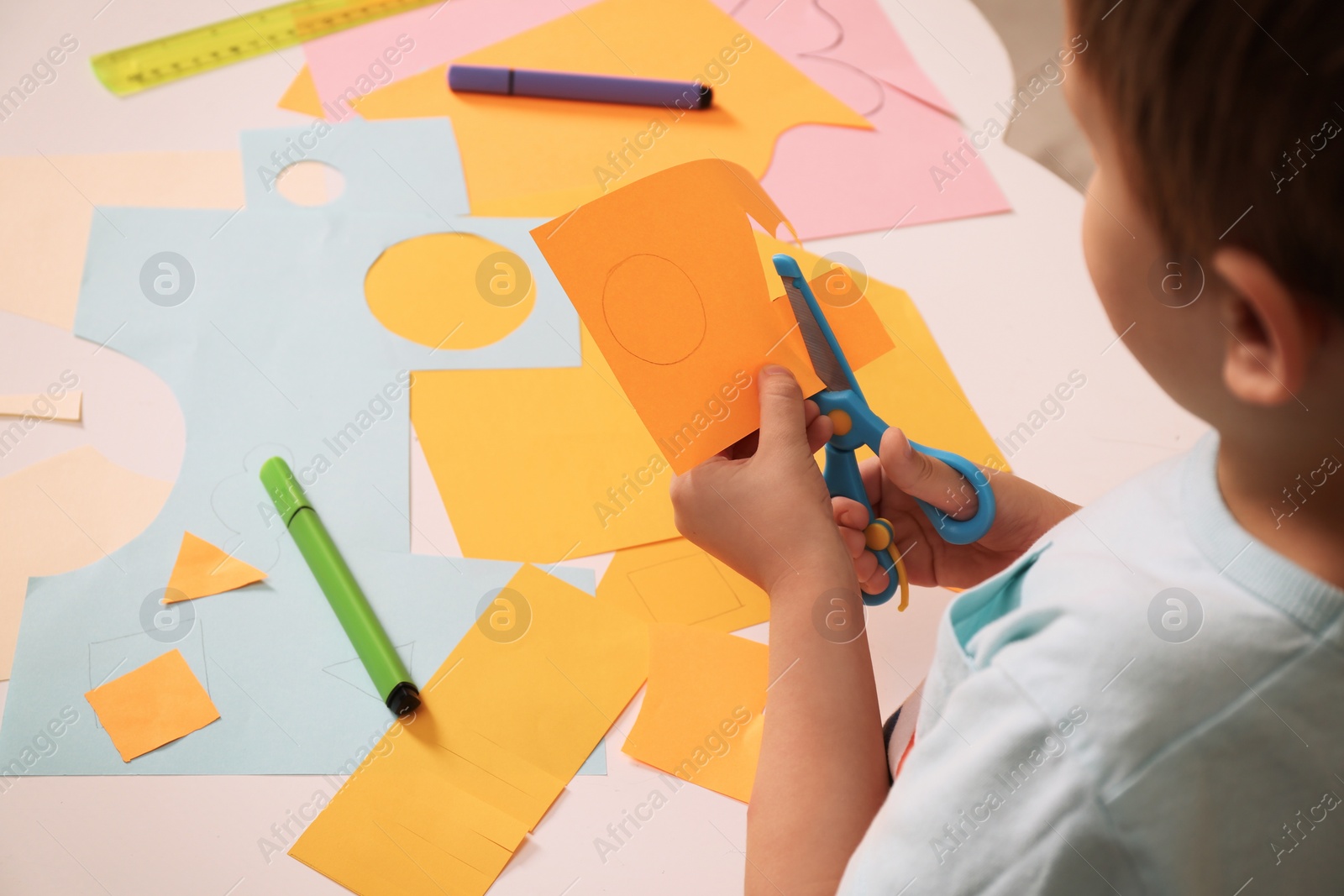 Photo of Little boy cutting color paper with scissors at table indoors, closeup