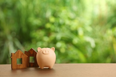 Piggy bank and house models on wooden table outdoors. Space for text