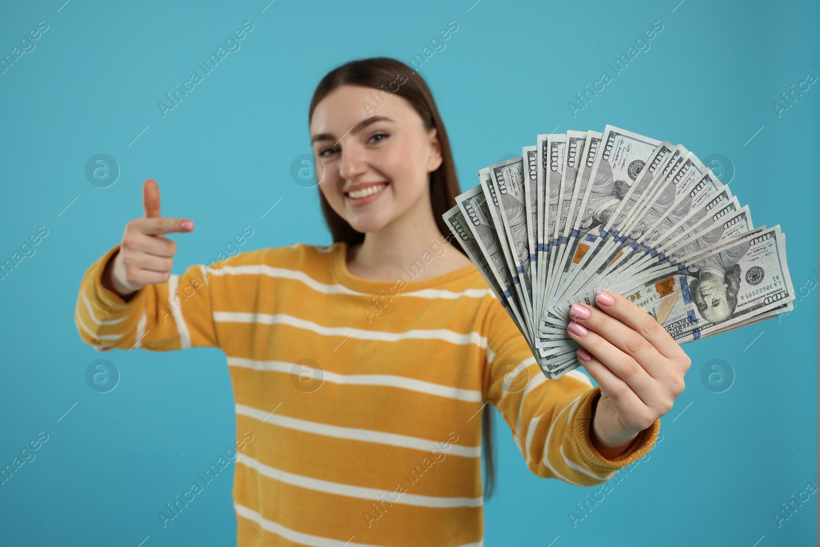 Photo of Happy woman pointing at dollar banknotes on light blue background, selective focus