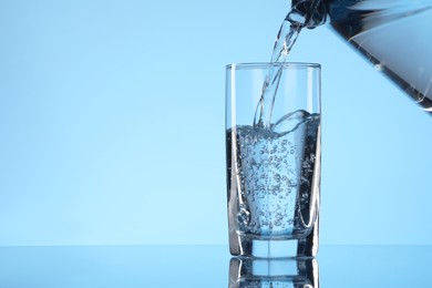 Pouring water from jug into glass on light blue background. Space for text