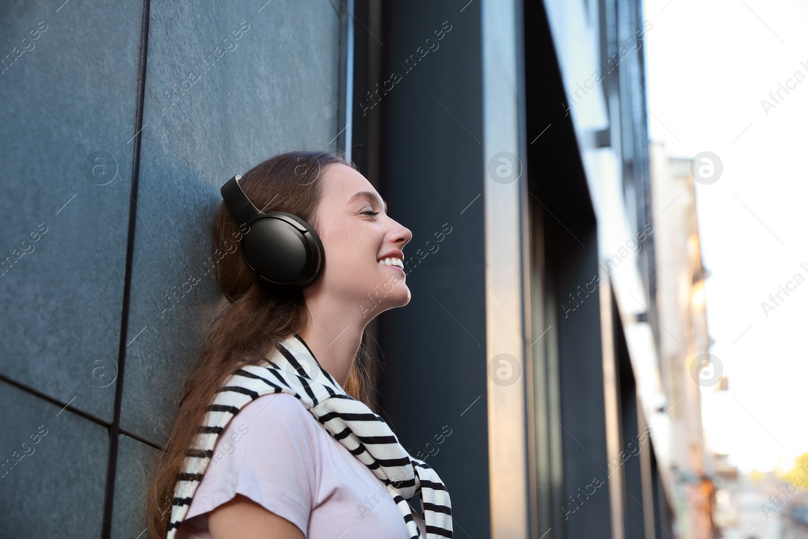 Photo of Smiling woman in headphones listening to music near building outdoors. Space for text