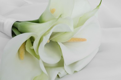 Photo of Beautiful calla lily flowers tied with ribbon on white fabric, closeup