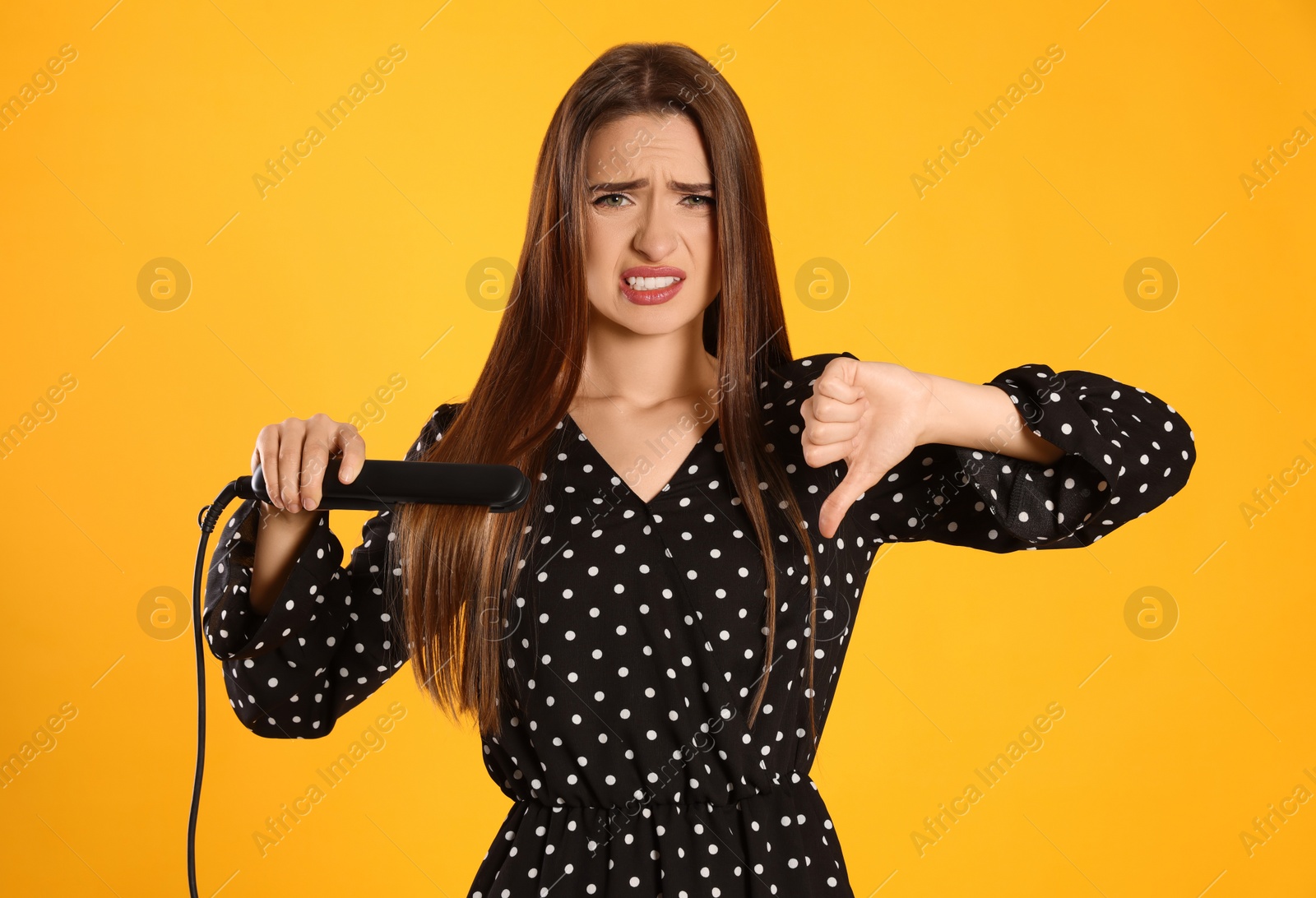 Photo of Upset young woman with flattening iron showing thumb down on yellow background. Hair damage