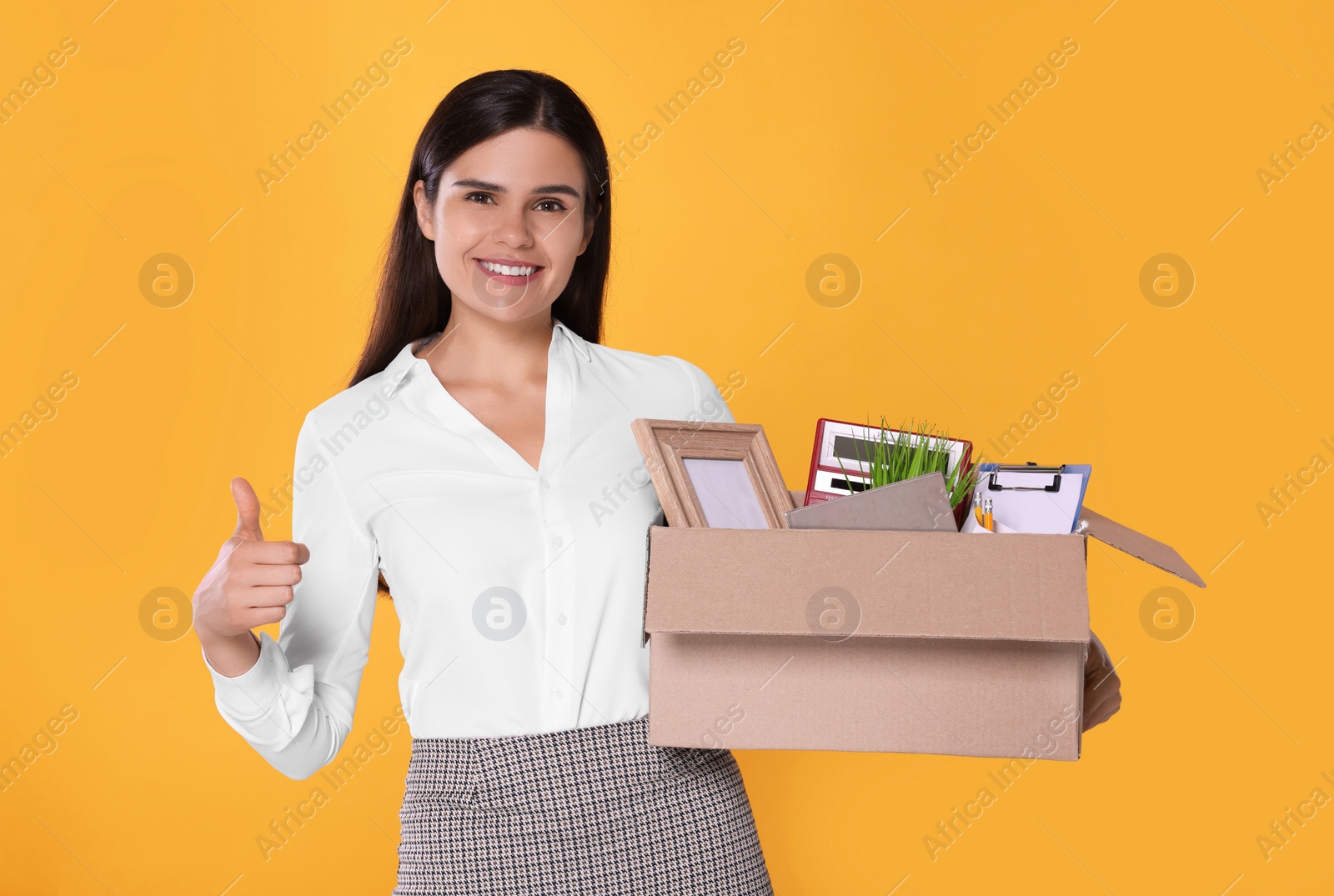 Photo of Happy unemployed woman with box of personal office belongings showing thumb up on orange background