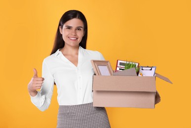 Photo of Happy unemployed woman with box of personal office belongings showing thumb up on orange background