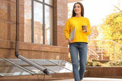 Image of Happy young woman with laptop and paper cup of coffee walking outdoors