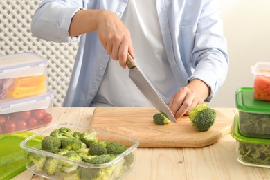 Photo of Man cutting fresh broccoli with knife near containers at wooden table, closeup. Food storage
