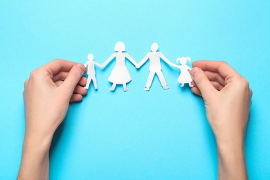 Woman holding paper cutout of family on light blue background, top view