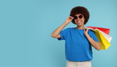 Happy young woman in stylish sunglasses with shopping bags on light blue background. Space for text