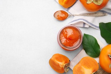 Jar and spoon of tasty persimmon jam, ingredients on white table, flat lay. Space for text