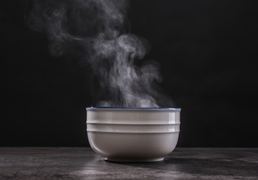 Photo of Steaming ceramic bowl on grey table against dark background