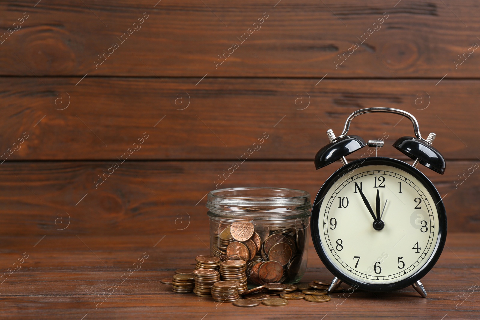 Photo of Black alarm clock, glass jar and coins on table against wooden background, space for text. Money savings