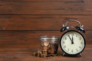 Black alarm clock, glass jar and coins on table against wooden background, space for text. Money savings