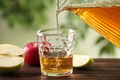 Pouring apple juice into glass on wooden table against blurred green background