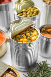 Photo of Open tin cans with corn kernels and different products on white table, closeup