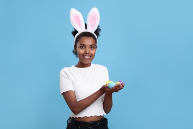 Photo of Happy African American woman in bunny ears headband holding Easter eggs on light blue background. Space for text