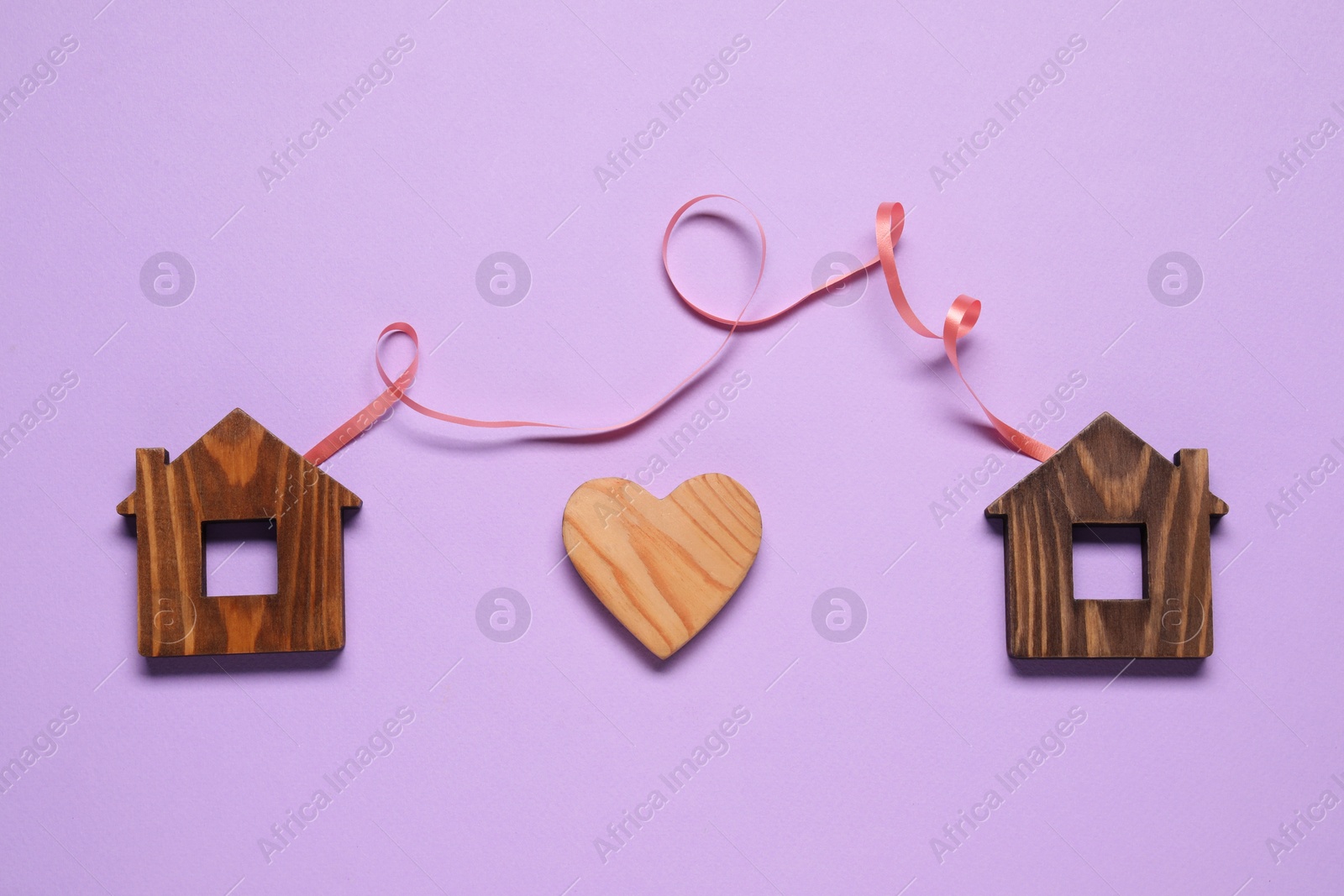 Photo of Ribbon and decorative heart between two wooden house models on violet background symbolizing connection in long-distance relationship, flat lay