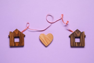 Ribbon and decorative heart between two wooden house models on violet background symbolizing connection in long-distance relationship, flat lay