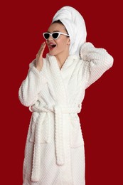 Beautiful young woman in bathrobe and sunglasses yawning on red background