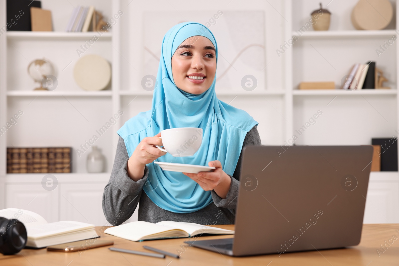 Photo of Muslim woman in hijab with cup of coffee using laptop at wooden table in room