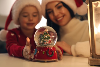 Photo of Mother and daughter in Santa hats with snow globe at table, focus on toy