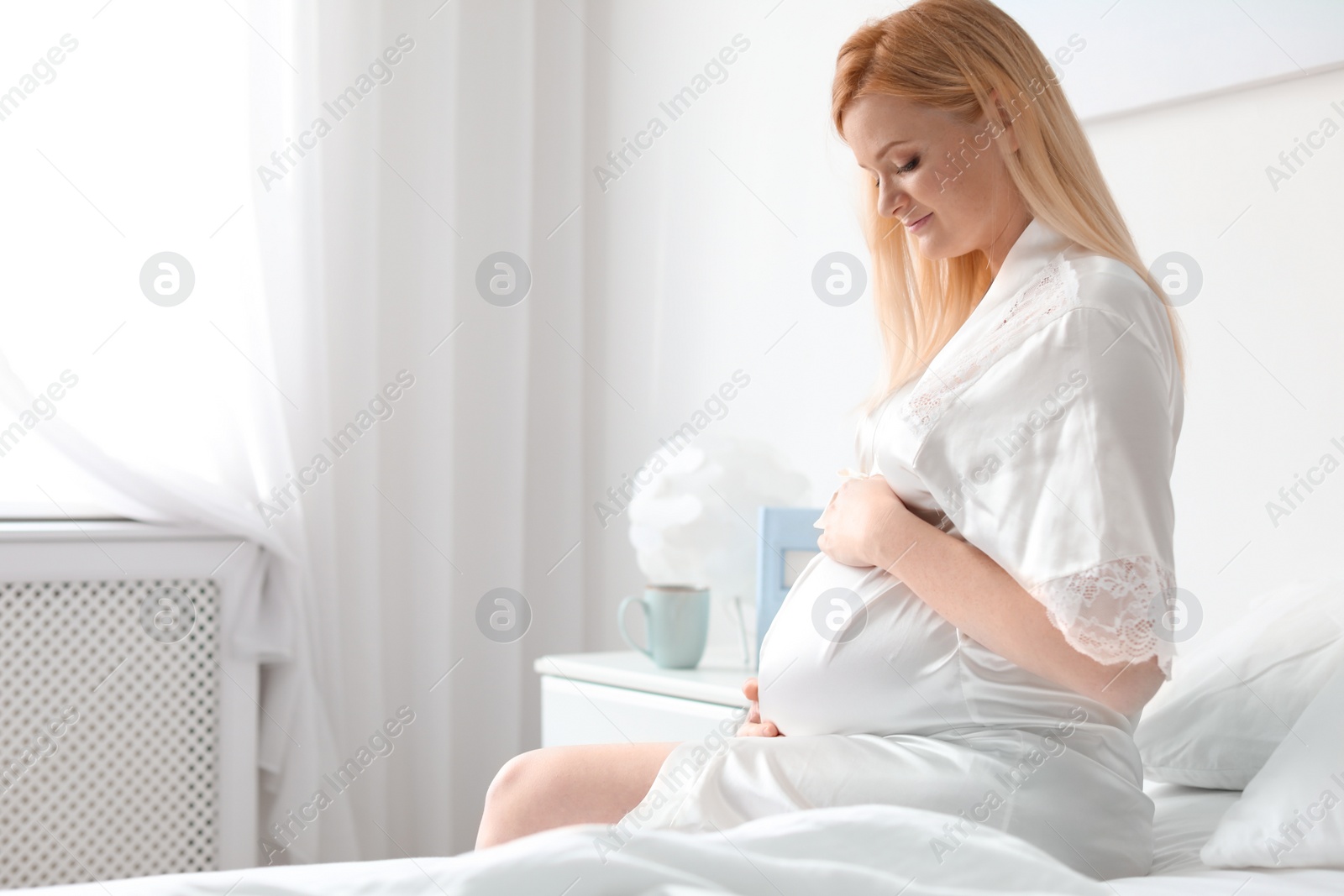 Photo of Beautiful pregnant woman sitting on bed in light room. Space for text