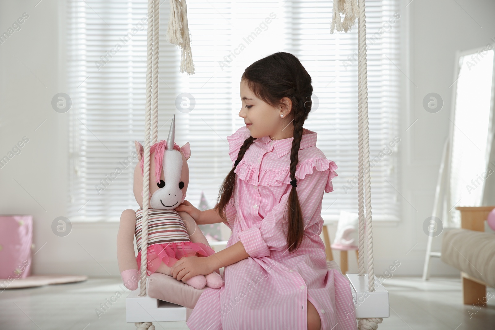 Photo of Cute little girl playing with toy unicorn on swing at home