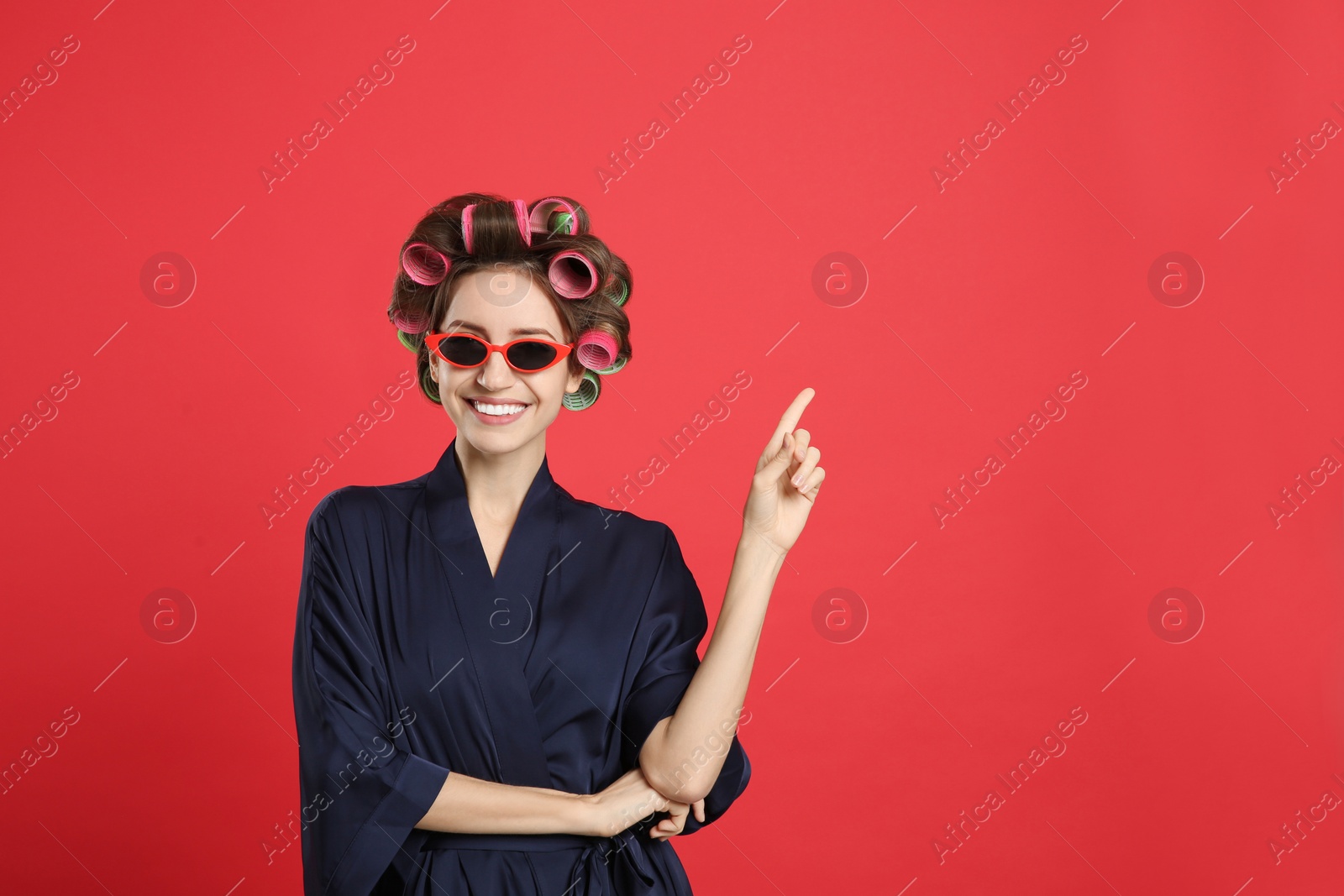 Photo of Beautiful young woman in silk bathrobe with hair curlers and sunglasses on red background