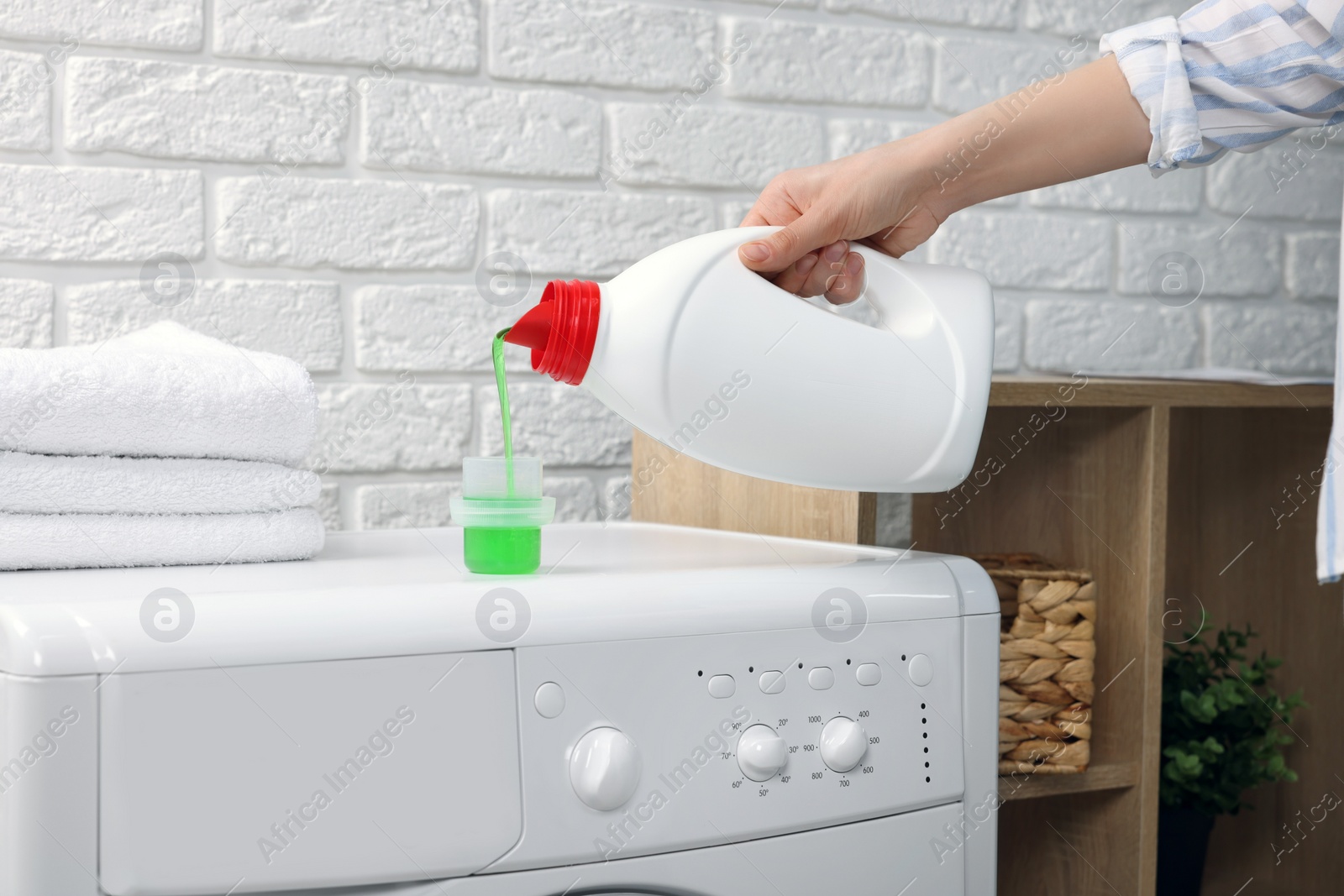 Photo of Woman pouring fabric softener from bottle into cap on washing machine indoors, closeup