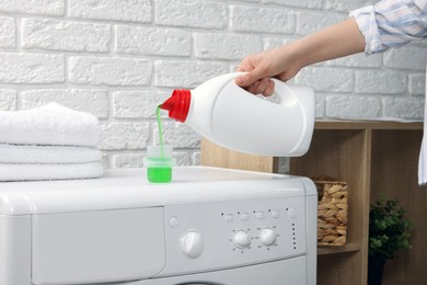 Photo of Woman pouring fabric softener from bottle into cap on washing machine indoors, closeup