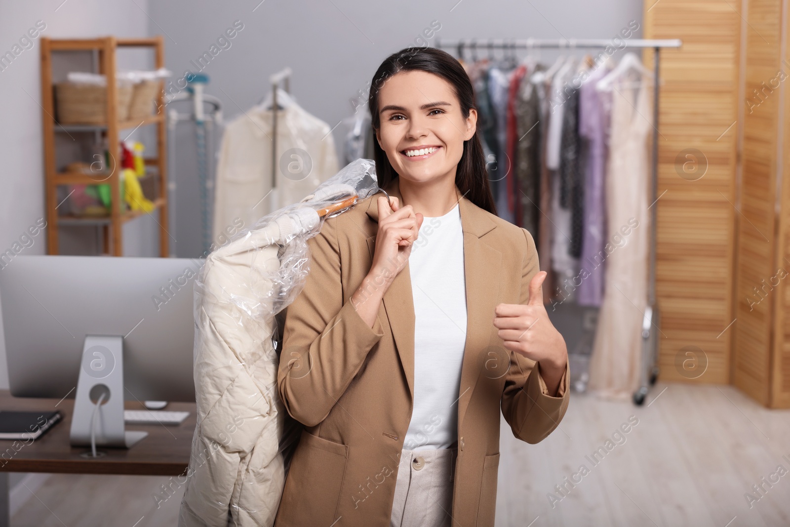 Photo of Dry-cleaning service. Happy woman holding hanger with jacket in plastic bag and showing thumb up indoors