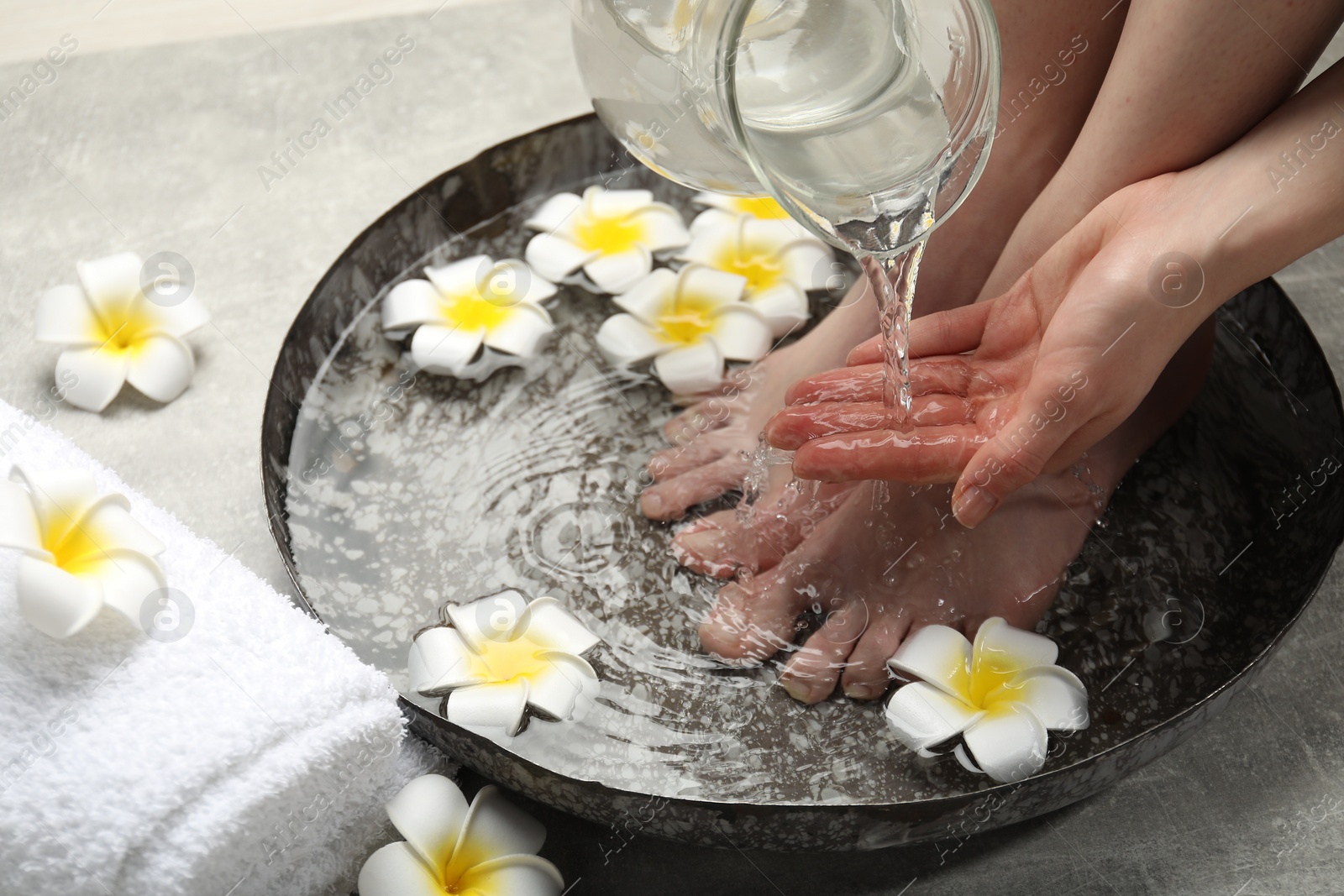 Photo of Woman pouring water onto hand while soaking her feet in bowl on light grey floor, closeup. Spa treatment