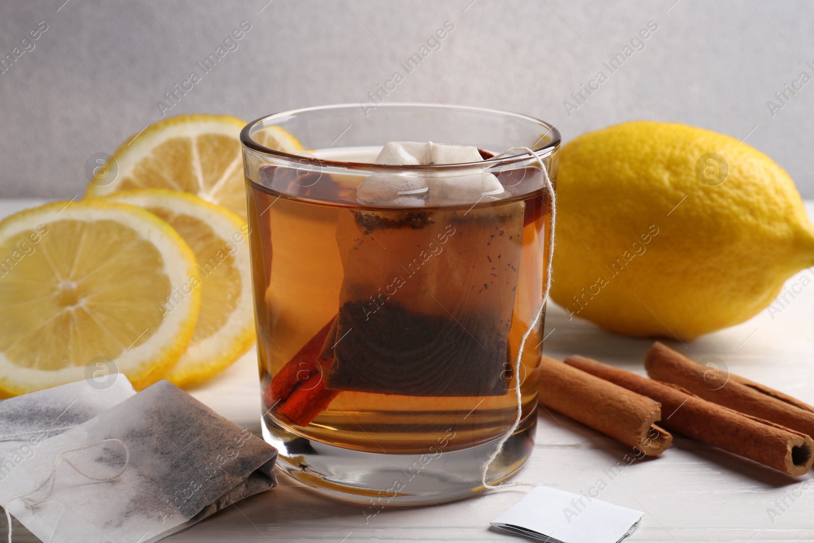 Photo of Tea bags, glass of hot drink, cinnamon sticks and lemons on white wooden table, closeup