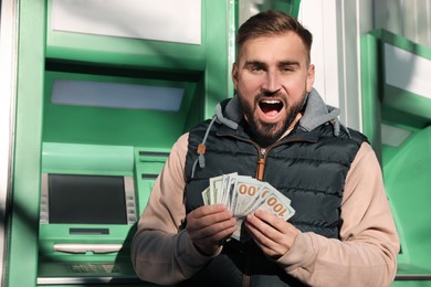 Excited young man with money near cash machine outdoors